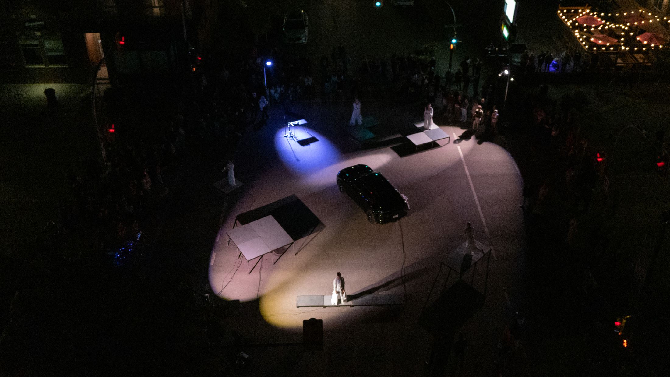 The intersection from above at night: a pool of white light around the car, raised platforms, performers in white, the audience at the edges