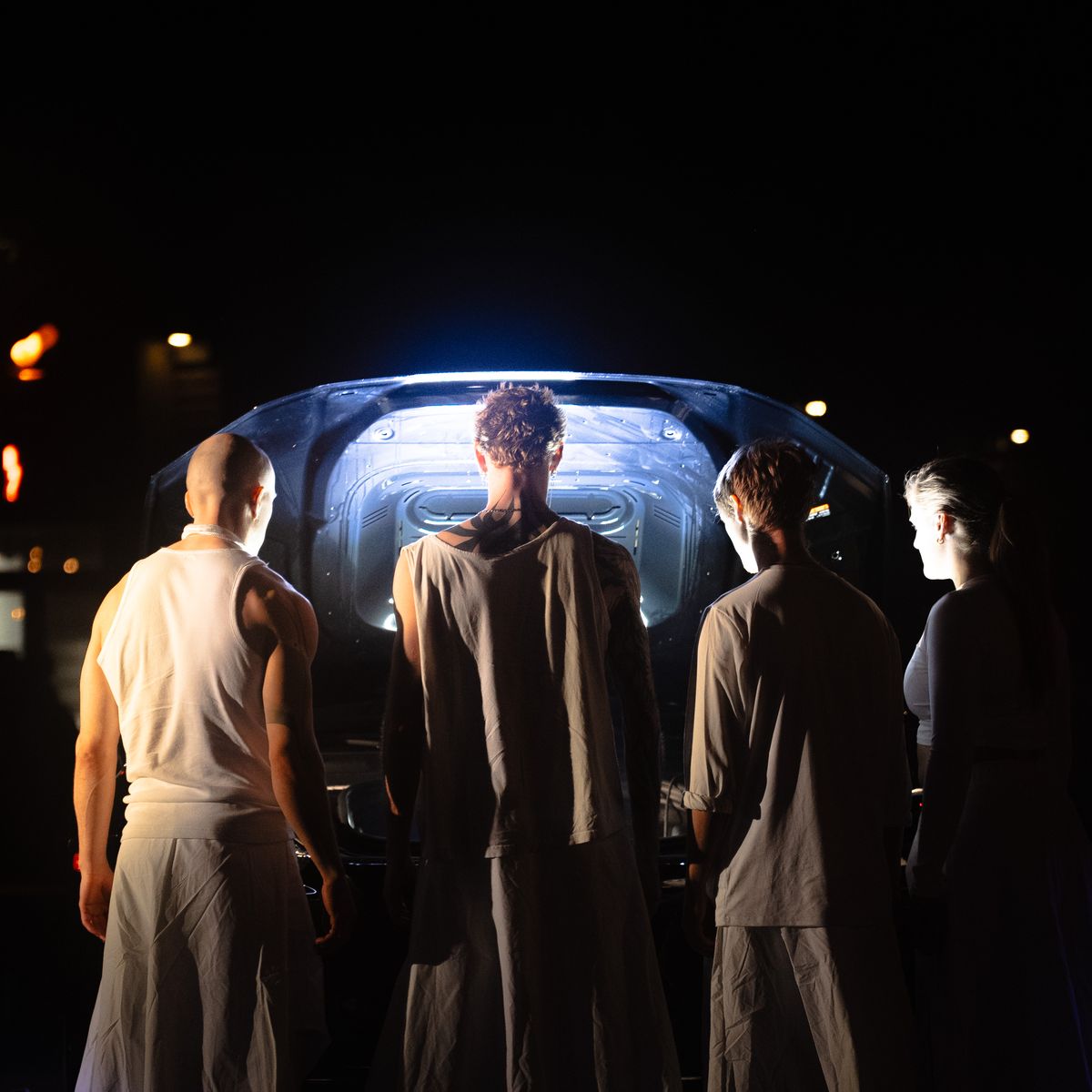 Performers in white gathered at the open back of the car, lit from inside it