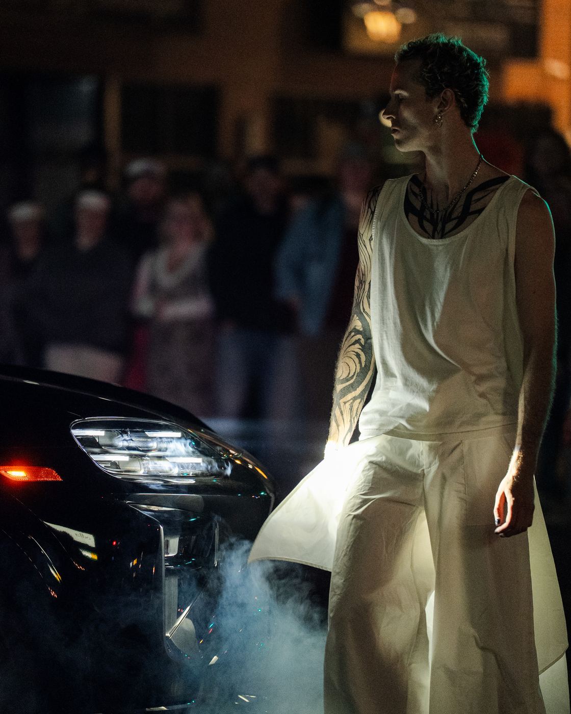 A dancer beside the car’s headlights in haze, the crowd behind
