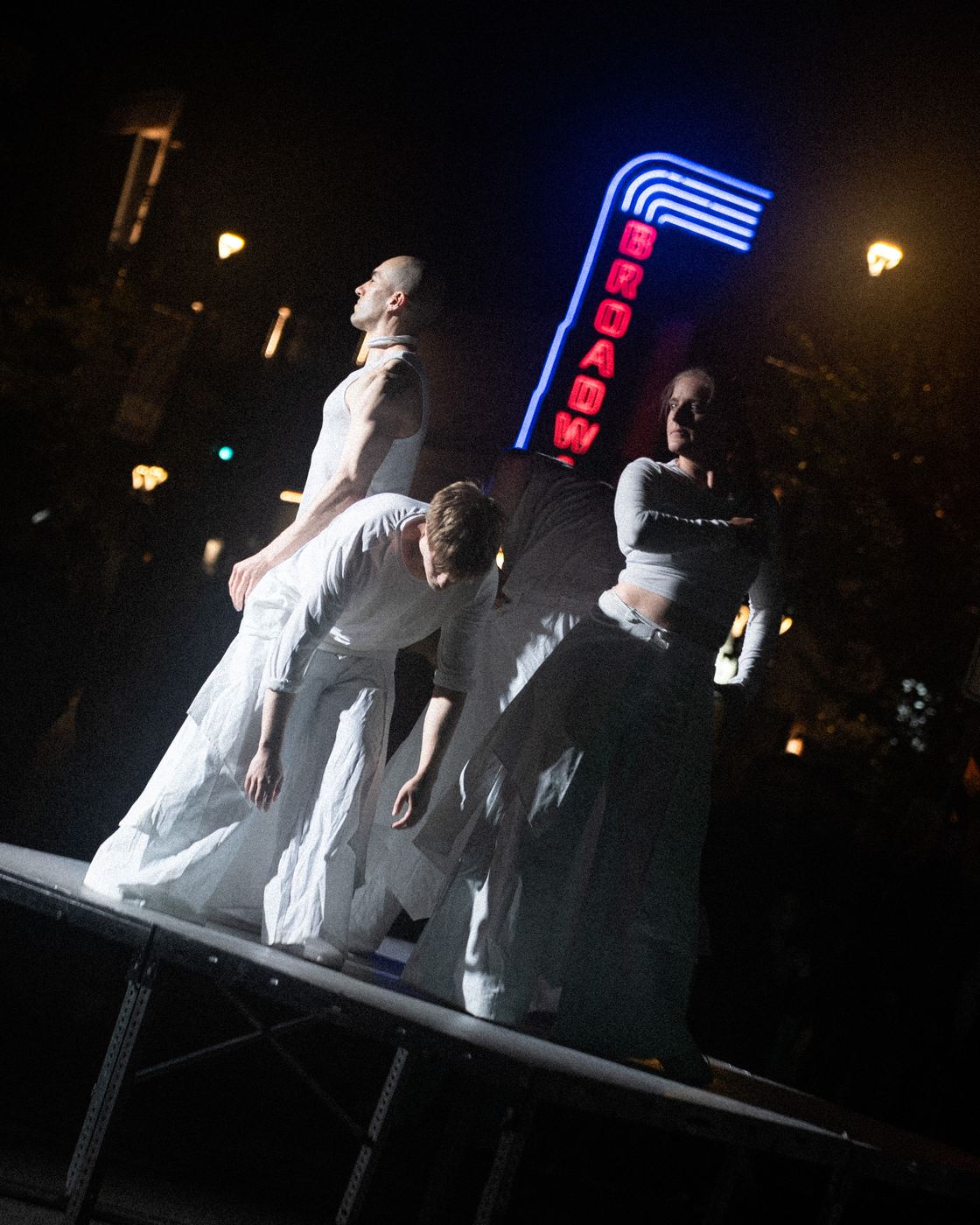 Three dancers on a tilted platform under the Broadway neon sign
